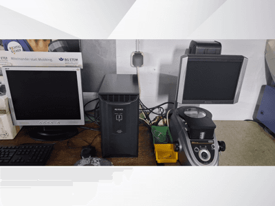 Two computer monitors, one Acer and one Keyence, next to a black device and a microscope, viewed from the front.